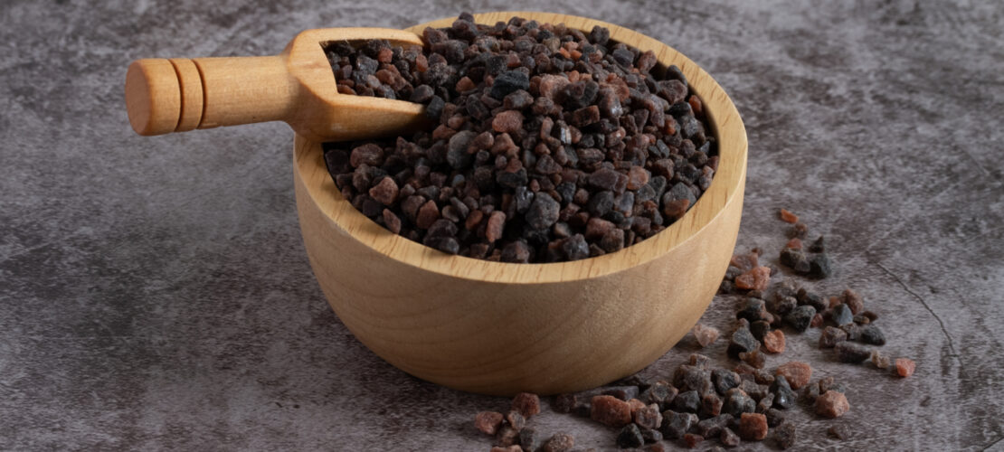 Eine Große Holzschüssel mit Holzlöffel auf grauem Untergrund. Die Schüssel ist mit Schwarzem Kala Namak Salz gefüllt