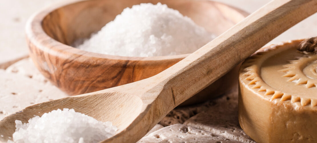 Eine Holzschüssel und Ein Holzlöffel liegen neben einem Stück seife auf Steinboden. Der Löffel und die Schüssel sind mit Badesalz Gefüllt